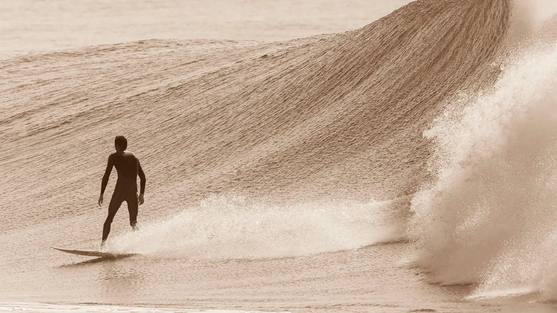 Surfer in a wetsuit riding a wave as a large swell breaks nearby, captured in sepia tone