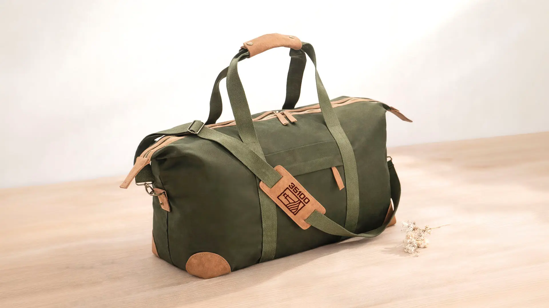 Green canvas duffel bag with tan leather accents and a branded 35100 luggage tag, displayed on a wooden surface