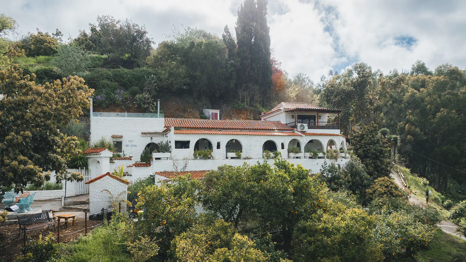 White Spanish colonial-style building with terracotta roof tiles and arched arcade, nestled among lush trees on a hillside
