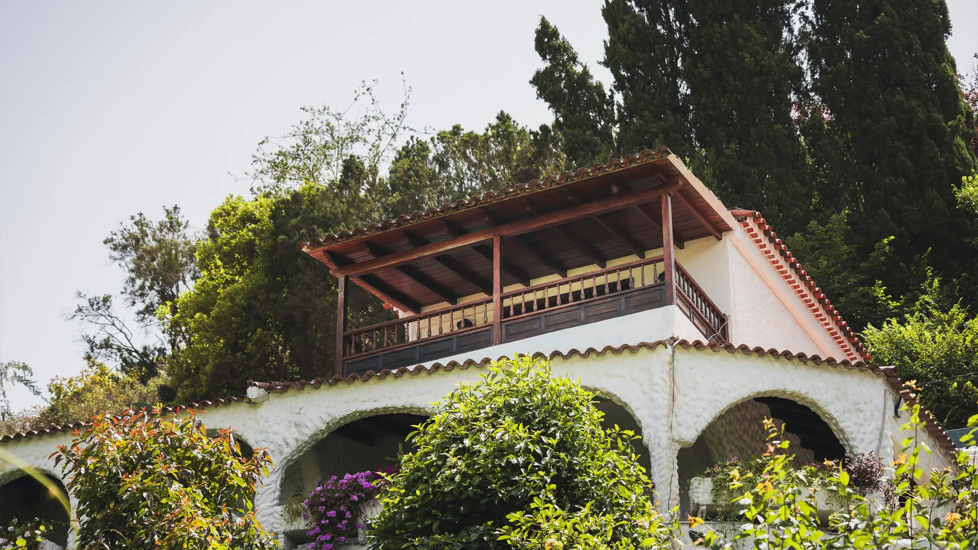 White stucco building with arched lower level and covered wooden balcony on upper floor, surrounded by lush greenery and flowering shrubs